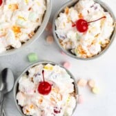 Three bowls of creamy Tropical Fluff fruit salad topped with maraschino cherries, surrounded by pastel mini marshmallows on a light surface, with two spoons placed nearby.