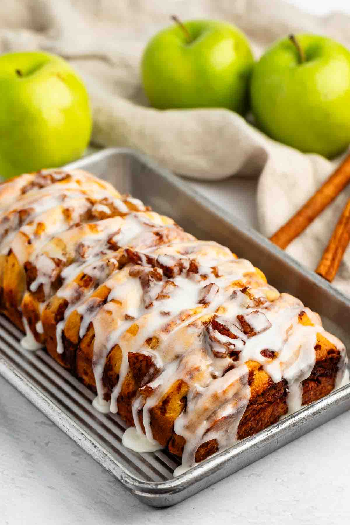 A glazed apple cinnamon bread loaf sits on a baking tray, topped with icing. In the background are two green apples, a beige cloth, and two cinnamon sticks.