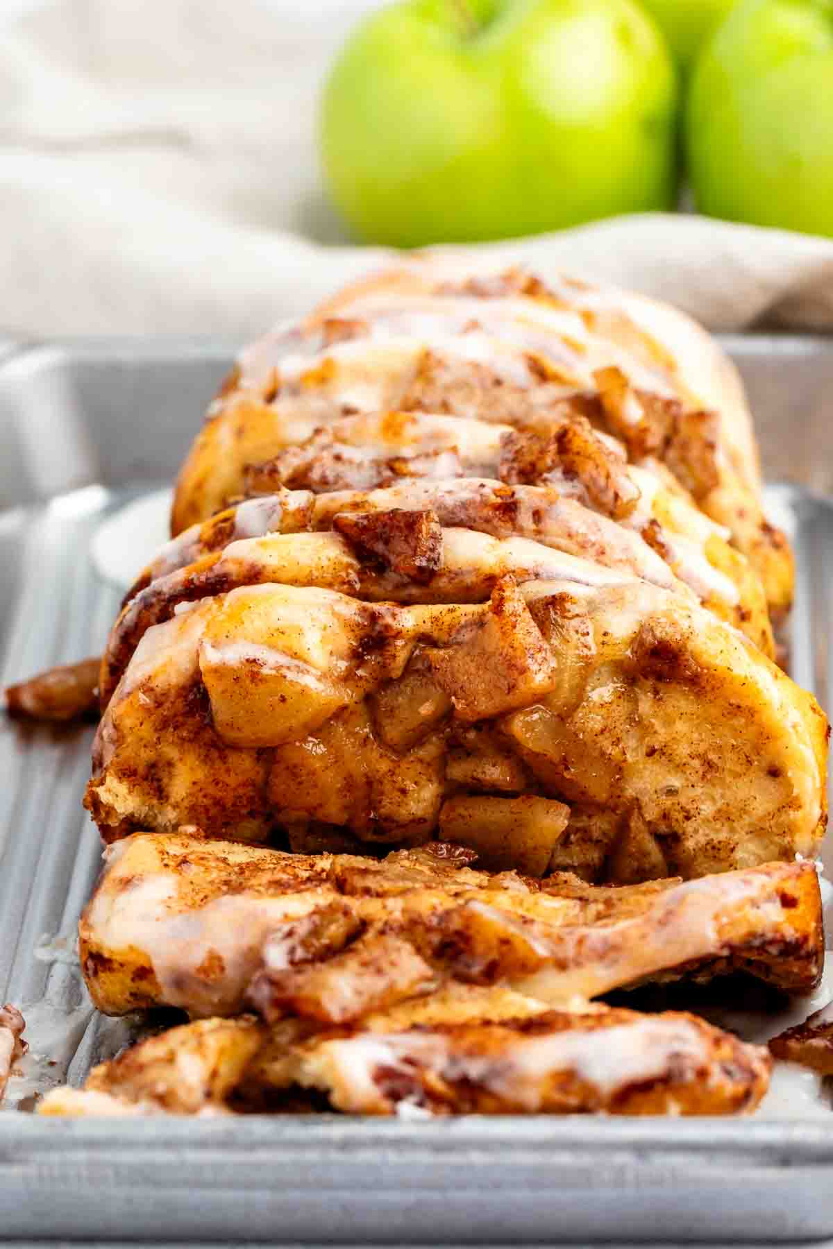 A close-up of sliced apple cinnamon bread topped with white icing, displayed on a metal tray with fresh green apples in the background.