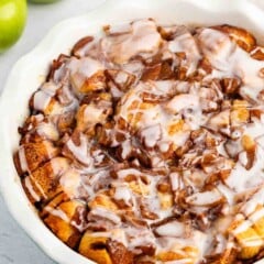 A baked apple cinnamon roll casserole in a white dish, topped with icing. Two green apples are in the background on a light surface.