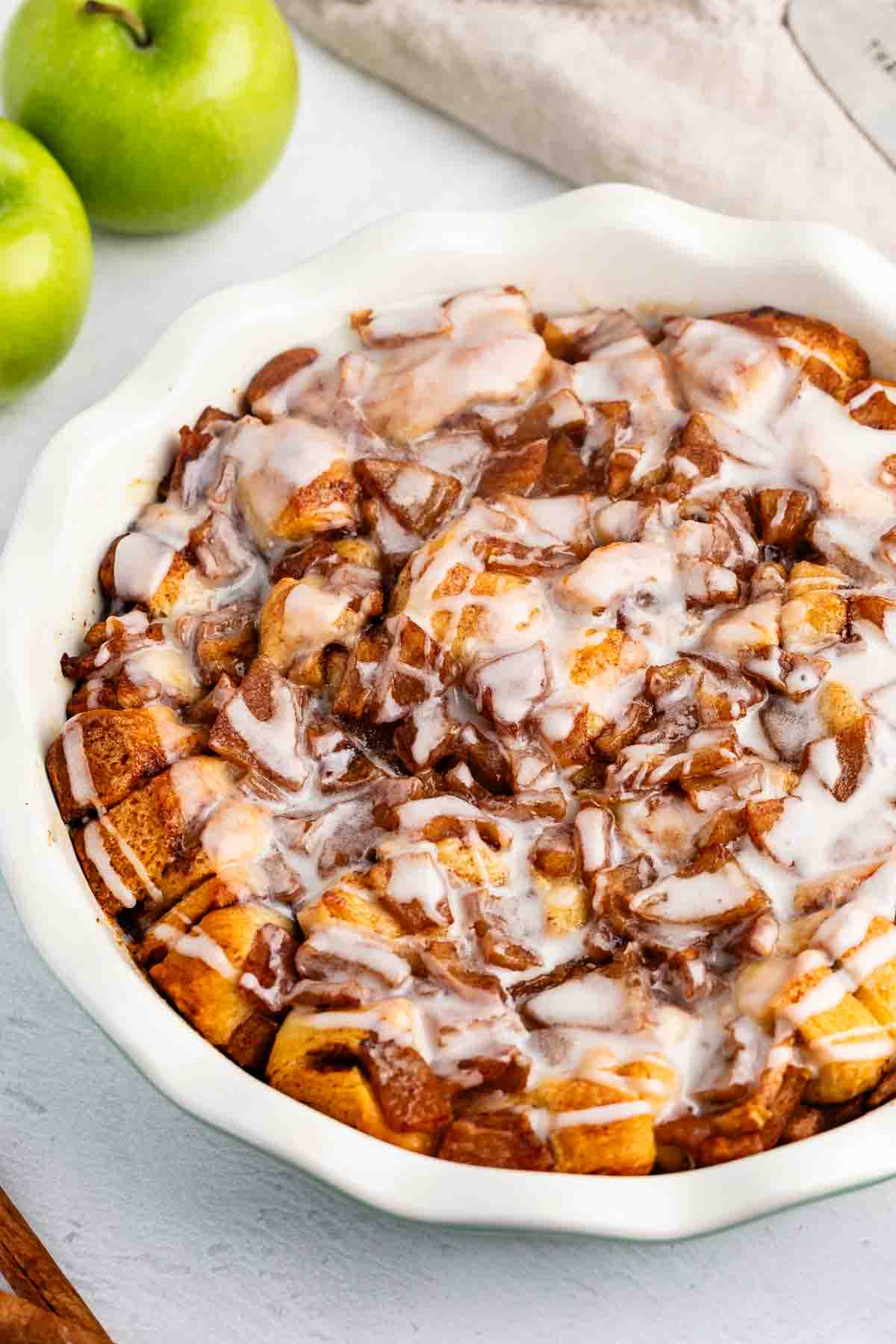 A baked apple cinnamon roll casserole in a white dish, topped with icing. Two green apples are in the background on a light surface.