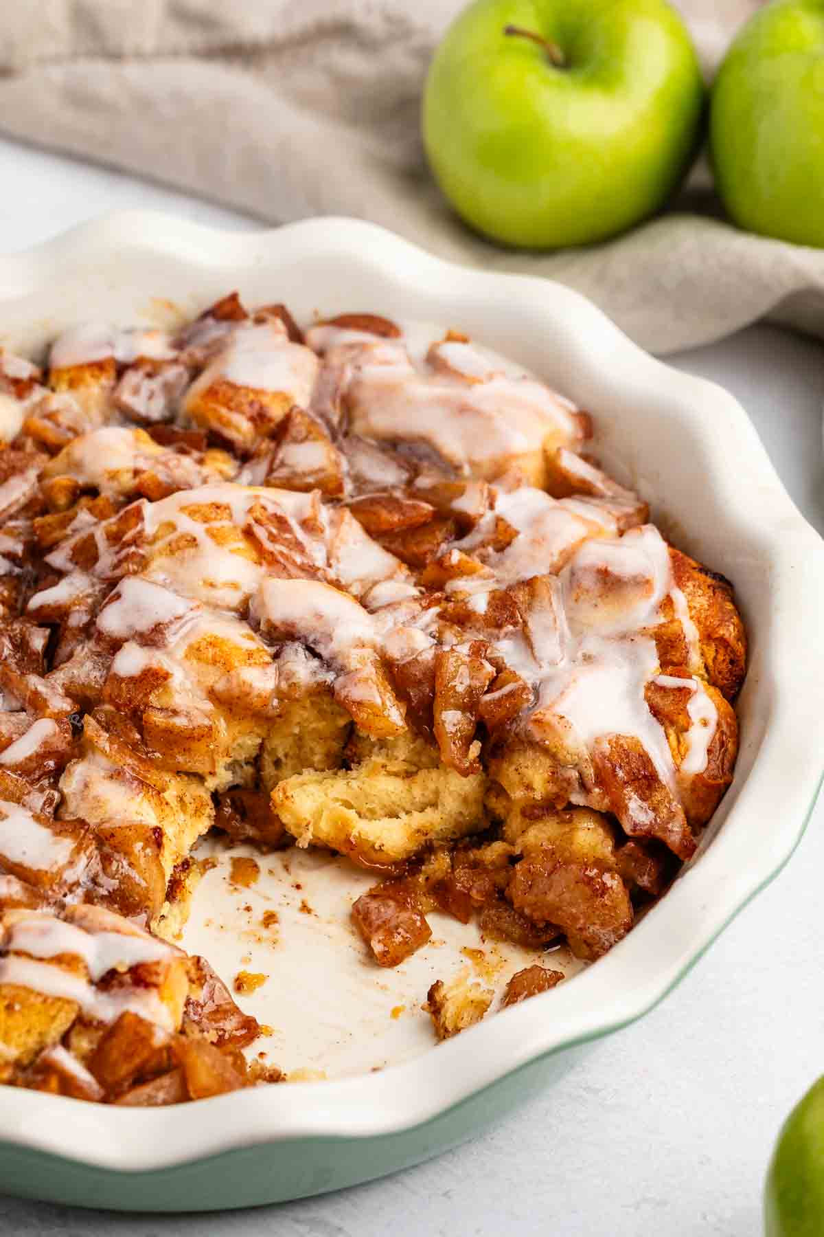 A baked cinnamon apple casserole topped with white icing in a round dish, with a portion removed. Two green apples and a beige cloth are in the background.