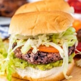 Veggie burger with a black bean patty, layered with fresh tomato slices, shredded lettuce, and guacamole, served in a soft bun on a wooden board. Blurred background shows additional ingredients and another burger.
