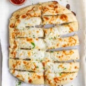 A rectangular cheese bread on a baking sheet lined with parchment paper, cut into strips. The bread is topped with melted cheese and garnished with parsley. A small cup of marinara sauce is on the side.