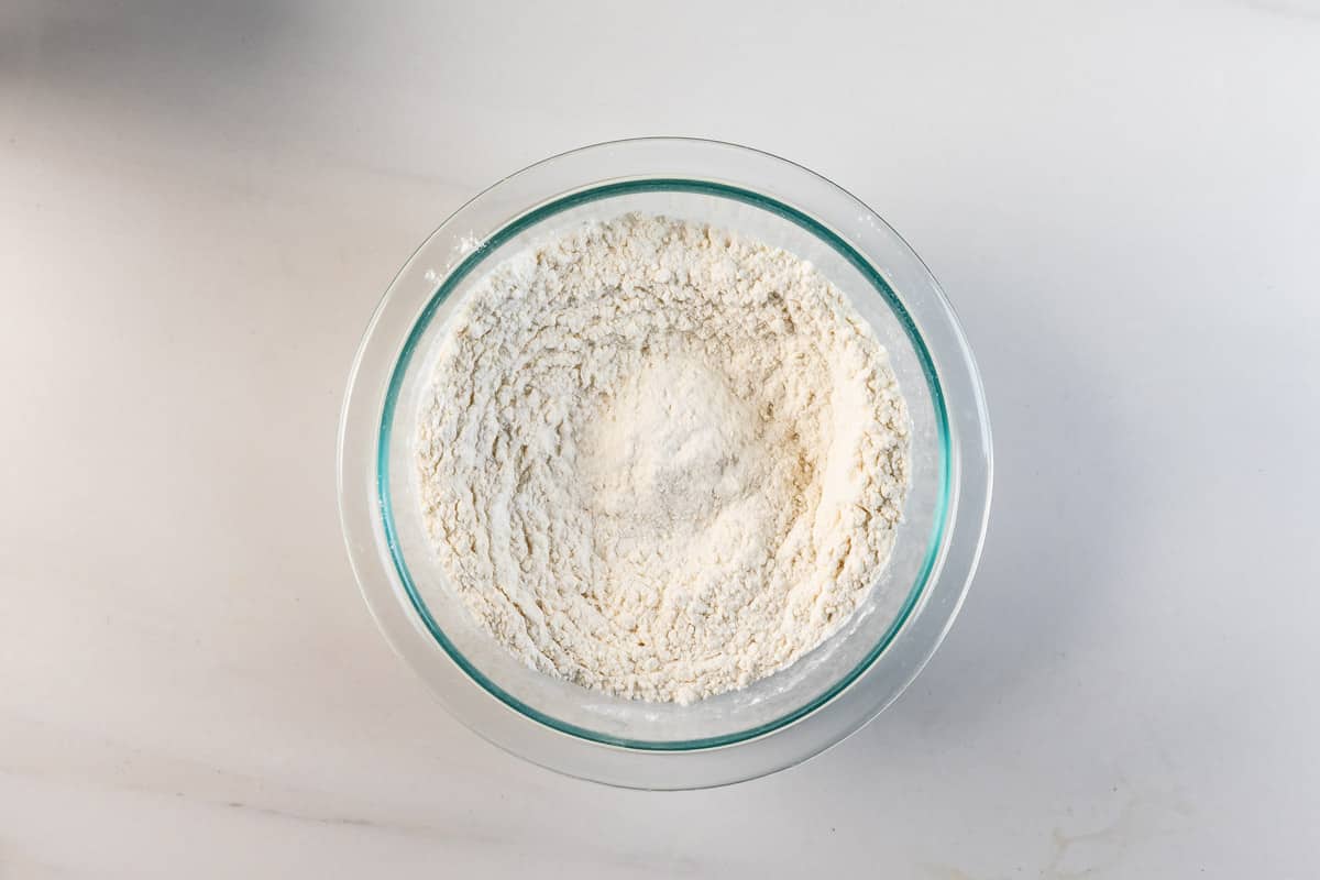 A glass bowl filled with flour sits on a white countertop. The flour is piled in the center, forming a small mound.