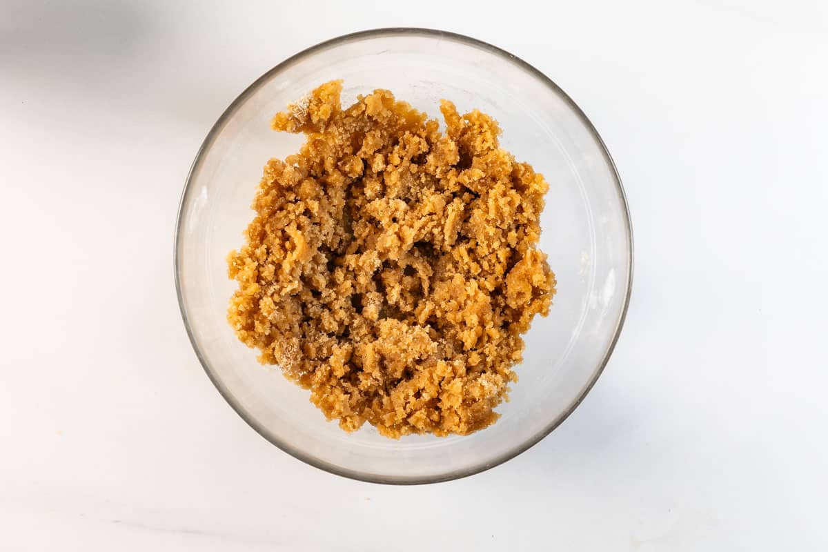 A clear glass bowl filled with a coarse, crumbly brown sugar mixture sits on a white surface.