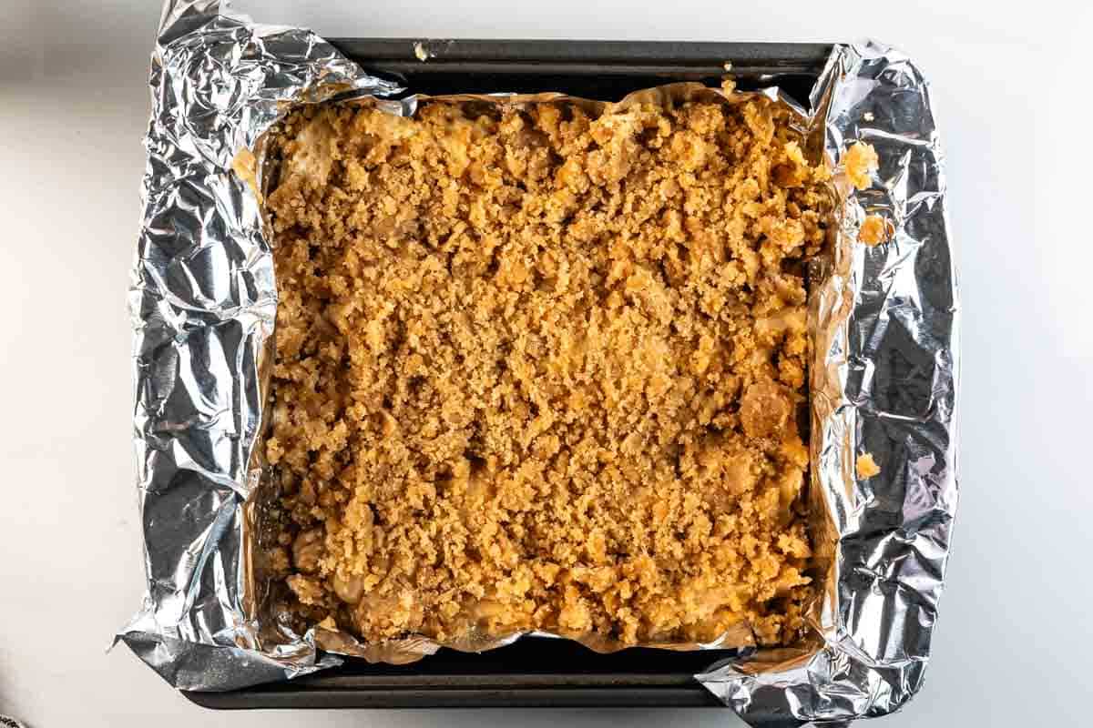 A square baking pan lined with crumpled aluminum foil, filled with an unbaked crumble mixture, viewed from above on a white surface.