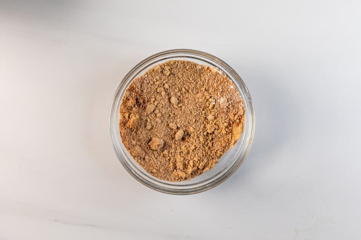 A glass bowl filled with light brown, finely crushed graham cracker crumbs sits on a white surface.
