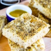 Two thick slices of focaccia bread stacked on a wooden board, topped with herbs and served with a small dish of olive oil for dipping. A bottle of wine is partially visible in the background.