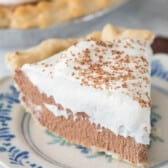 A slice of rich chocolate cream pie topped with whipped cream and chocolate shavings sits on a decorative plate, while the rest of the chocolate cream pie is visible in the background.