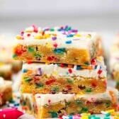Three funfetti cookie bars stacked on each other, topped with white frosting and colorful sprinkles. More cookie bars and scattered sprinkles can be seen in the background.