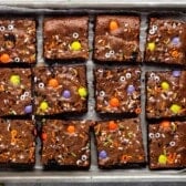 A tray of twelve halloween brownies topped with colorful Halloween-themed sprinkles, candy eyes, and orange, yellow, and purple candy-coated chocolates. The brownies are arranged neatly on parchment paper.