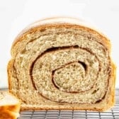 A close-up of a loaf of cinnamon swirl bread, sliced to show the spiral pattern inside, resting on a wire cooling rack with a light background.