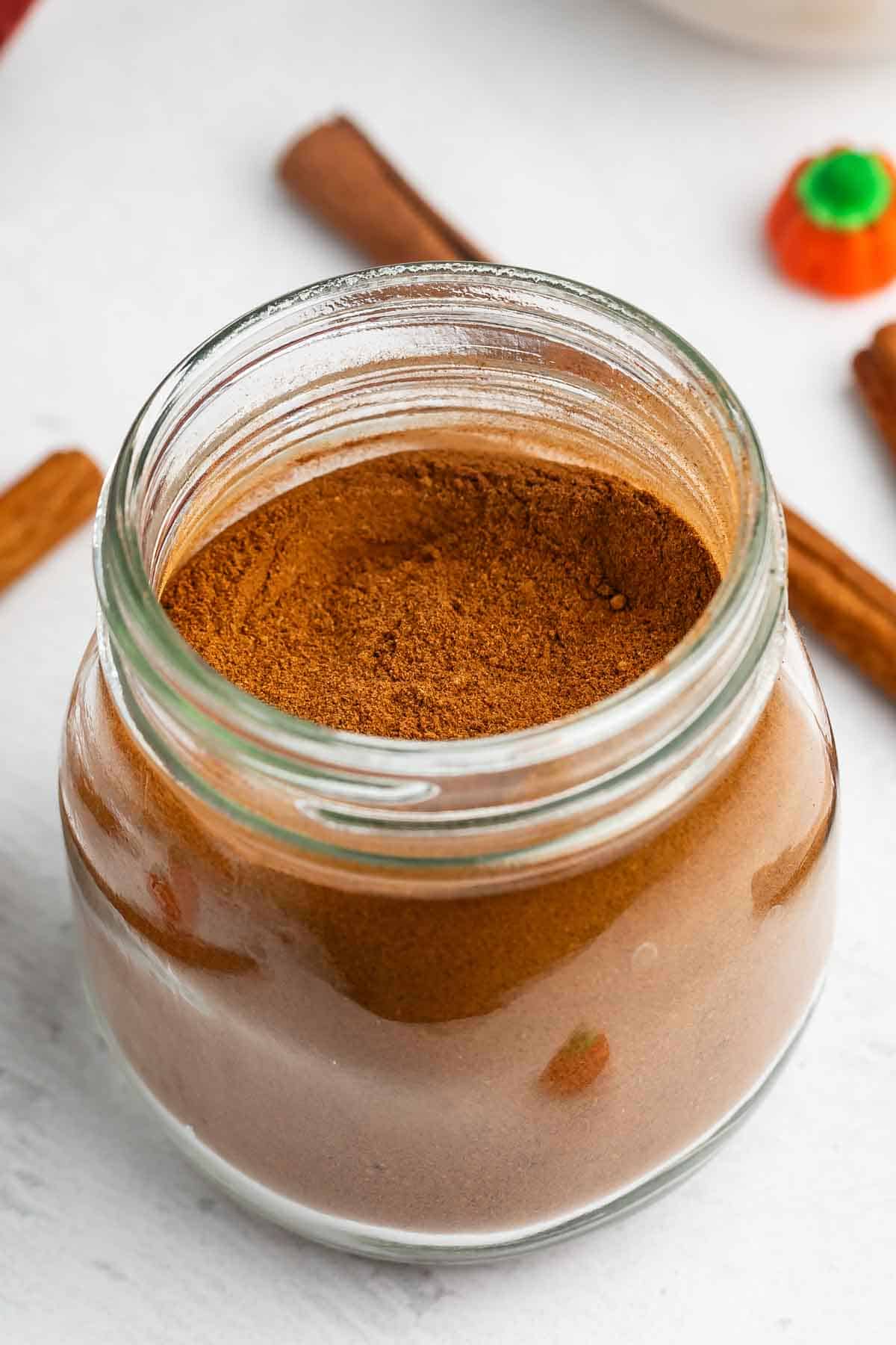 A glass jar filled with ground cinnamon sits on a white surface, with cinnamon sticks and a small pumpkin decoration nearby.