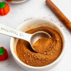 A white bowl filled with ground pumpkin spice, a metal measuring spoon resting inside. Cinnamon sticks and small pumpkin decorations are nearby on a white surface.