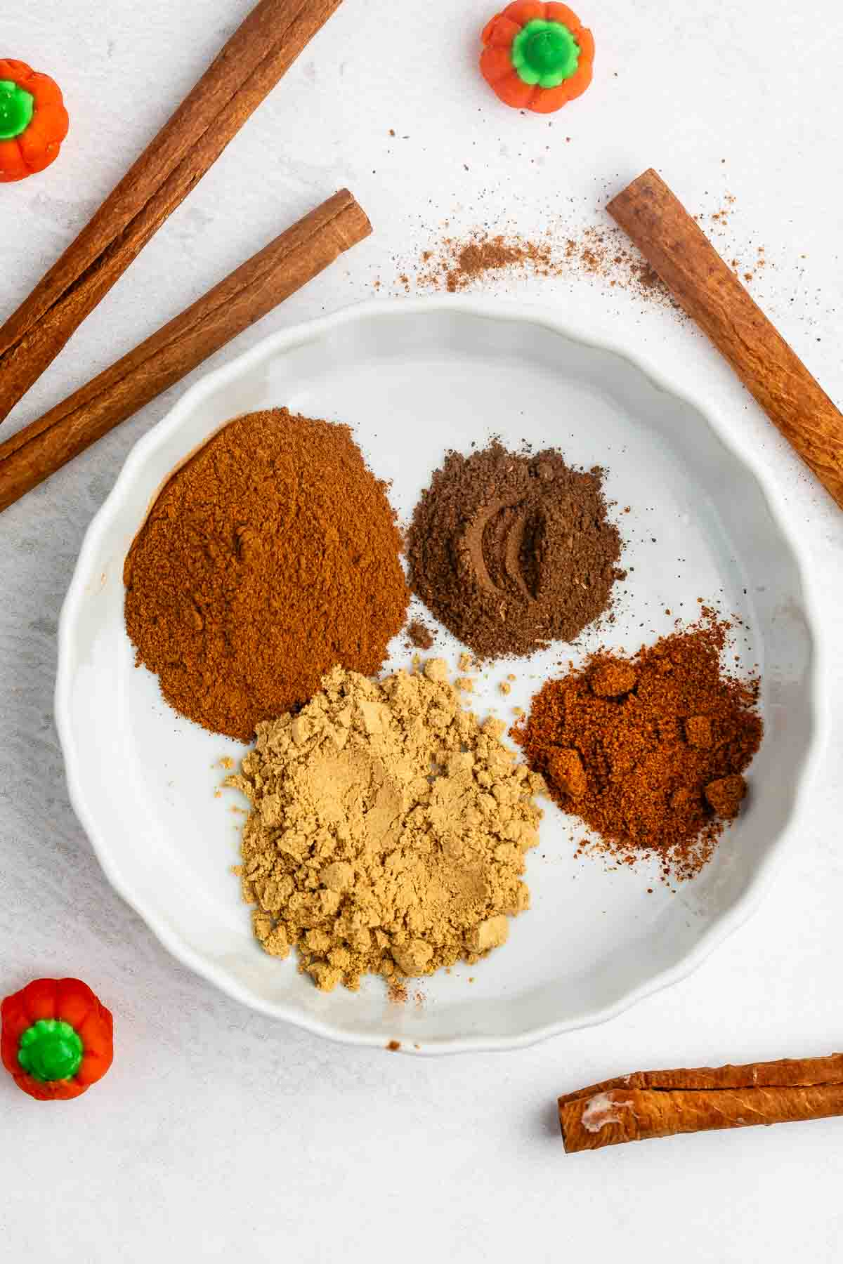 A white plate with four piles of ground spices—cinnamon, ginger, nutmeg, and cloves—surrounded by whole cinnamon sticks and small pumpkin-shaped decorations on a light surface.