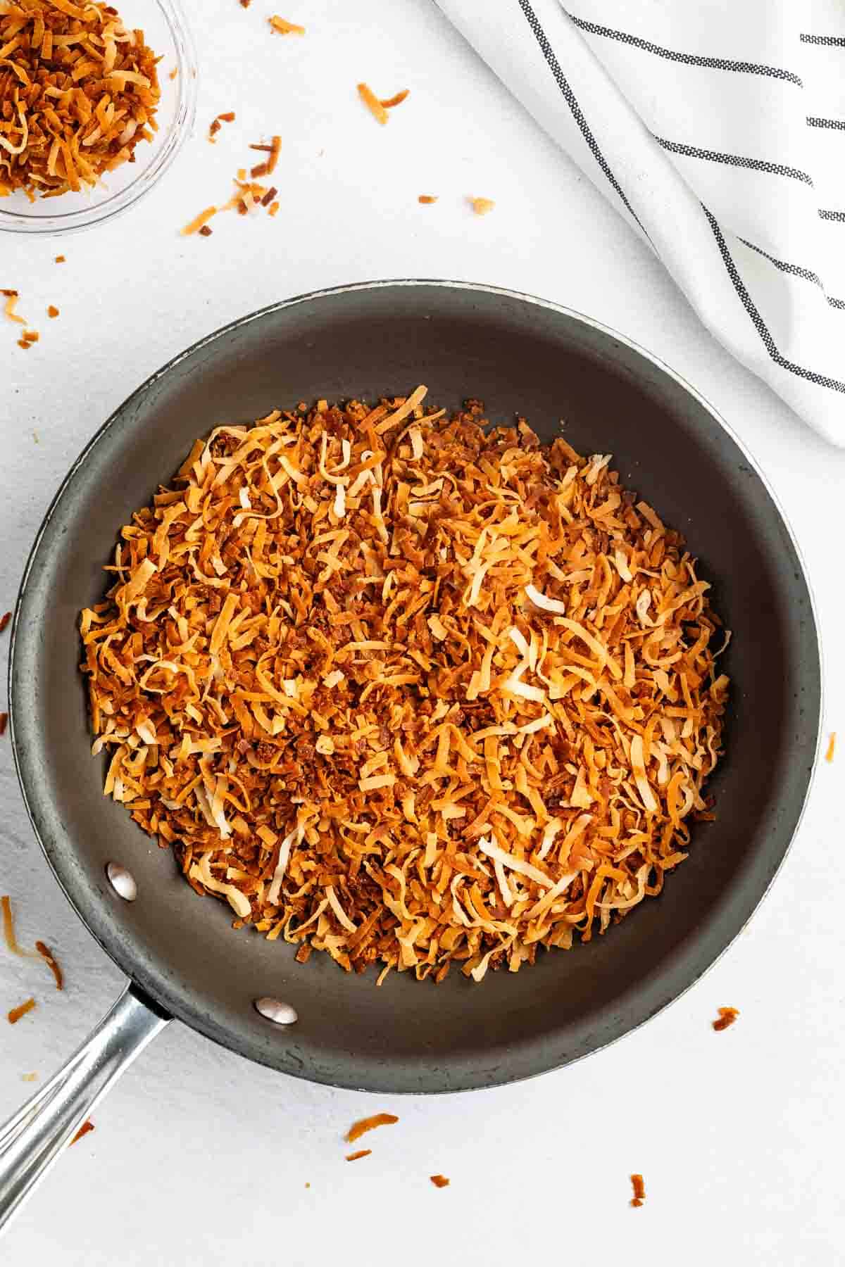 A grey pan filled with golden-brown toasted shredded coconut sits on a white surface, with a striped cloth in the background and a few coconut flakes scattered nearby.