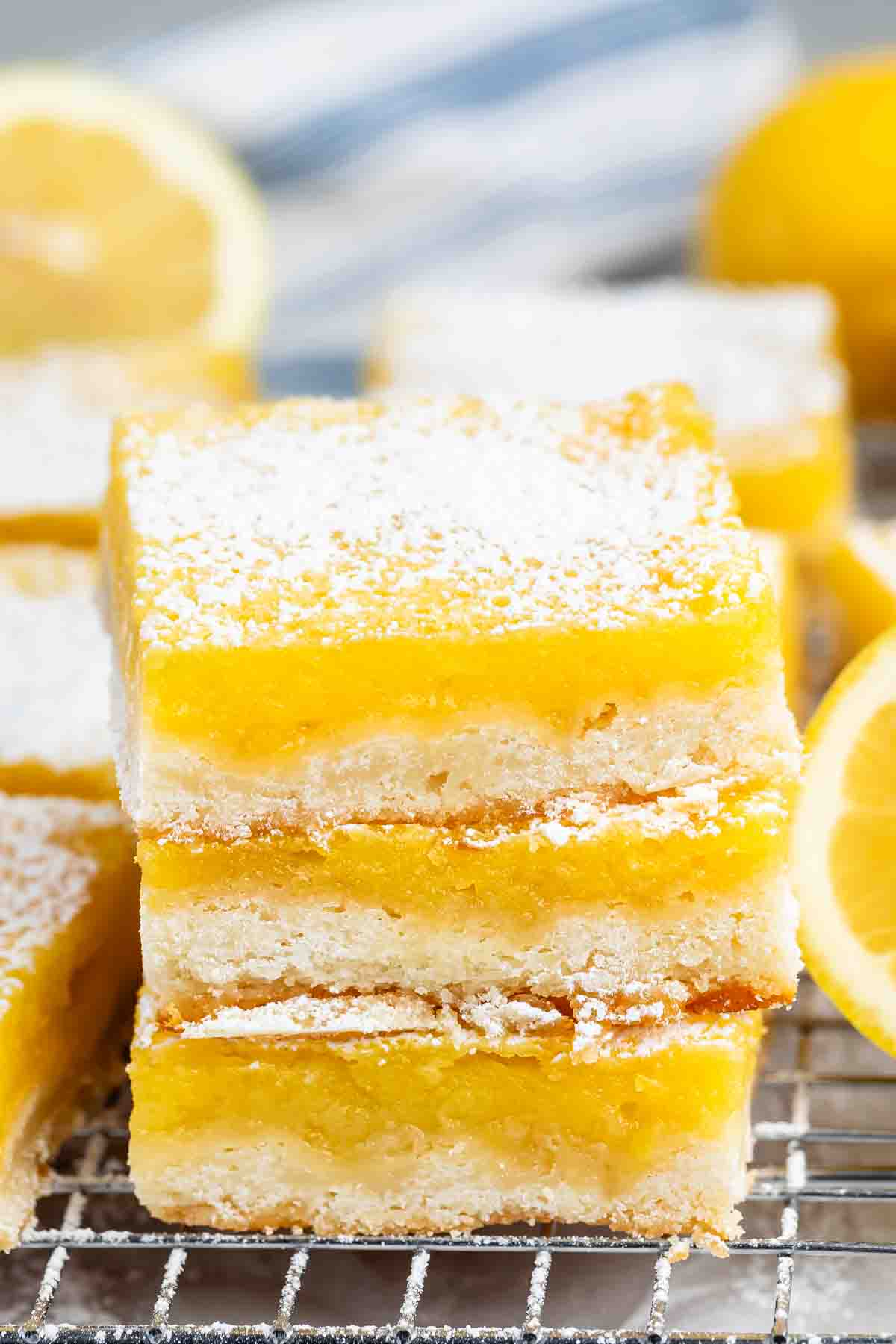 Close-up of lemon bars on a cooling rack, topped with powdered sugar. A fresh lemon is blurred in the background.