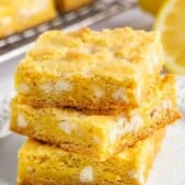 Three square lemon bars stacked on a white plate, with a bright yellow color and visible chunks, set in front of a wire rack and a sliced lemon in the background.