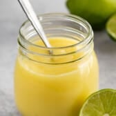 A glass jar filled with creamy yellow lime curd, with a metal spoon inside. In the background, there are whole and halved limes on a light gray surface.