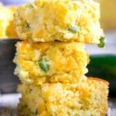 Three pieces of jalapeño cheddar cornbread stacked vertically. The bread appears moist with visible chunks of cheddar and bits of jalapeño, set on a rustic wooden surface with scattered shredded cheese and a blurred background.