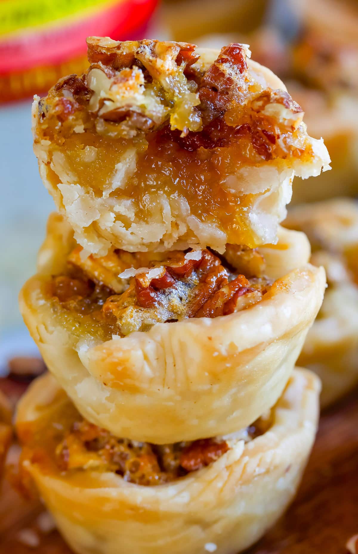 A close-up of three stacked mini pecan pies with flaky golden crusts and gooey, caramelized pecan filling, the top tart partially bitten to reveal the sticky interior.