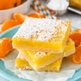 Three lemon bars stacked on a light blue plate, dusted with powdered sugar. In the background, there are sliced oranges, striped straws, and a small bowl of sugar on a white surface.