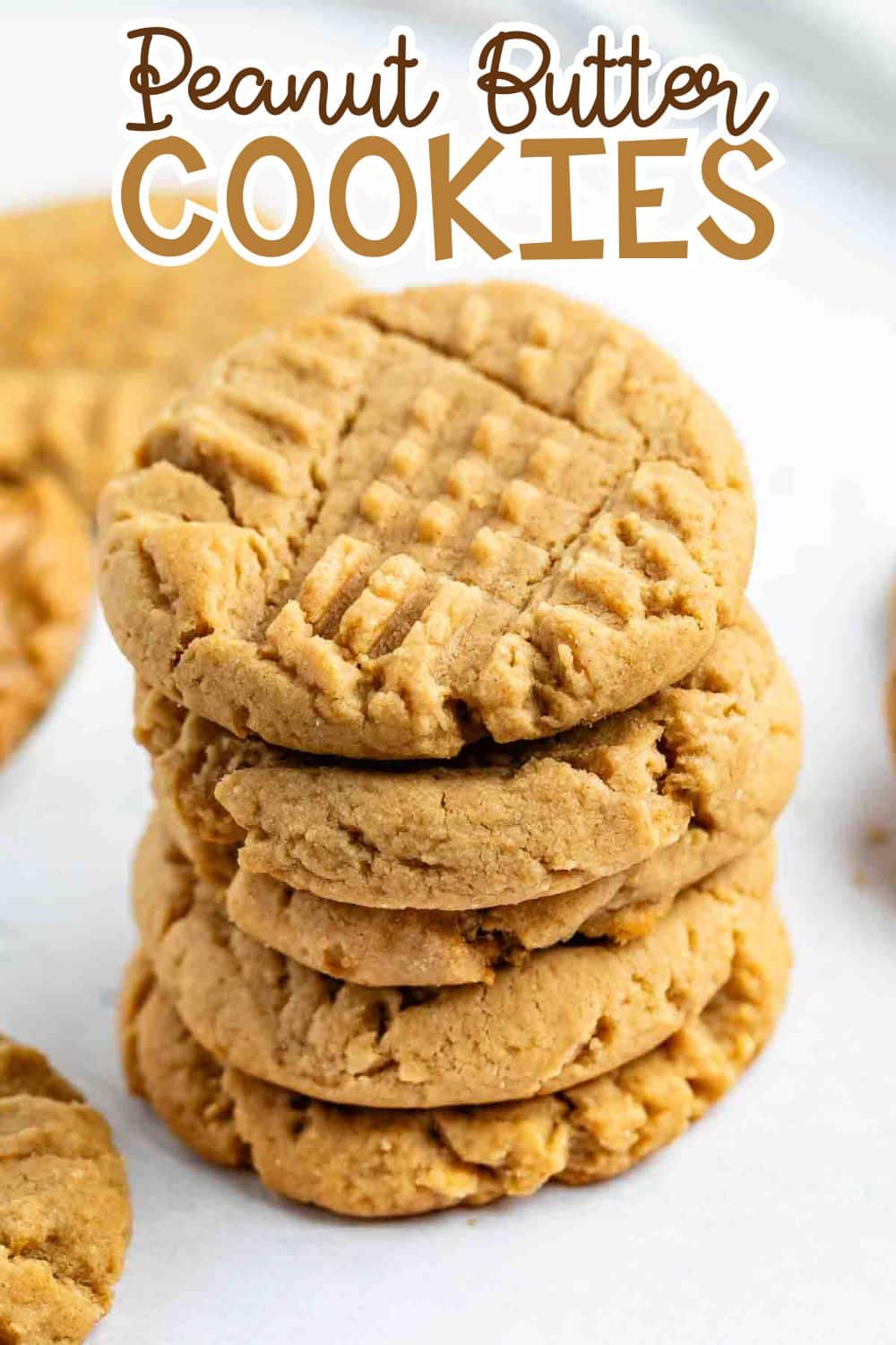 A stack of homemade peanut butter cookies with a crisscross fork pattern on top, surrounded by more cookies on a white surface. The text Peanut Butter Cookies is written above in playful fonts.