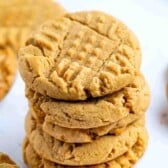 A stack of four golden brown peanut butter cookies with a crisscross fork pattern on top sits on a white surface, tempting anyone searching for the perfect peanut butter cookies recipe. More cookies are blurred in the background.