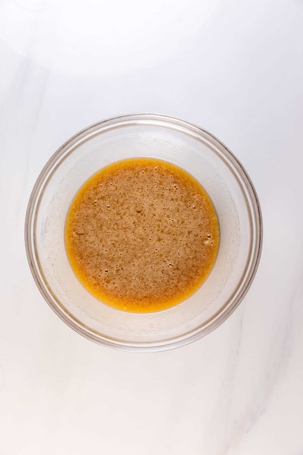 A glass bowl containing a light brown, frothy liquid mixture sits on a white marble surface.