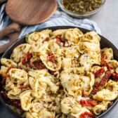 A bowl of tortellini pasta is topped with pesto, sun-dried tomatoes, and pine nuts. In the background, theres a small bowl of extra pesto, a bowl of grated cheese, and wooden serving utensils on a striped cloth.