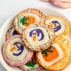 A plate of colorful Halloween cookies decorated with shapes of pumpkins and ghosts, surrounded by festive orange, purple, and black sprinkles.