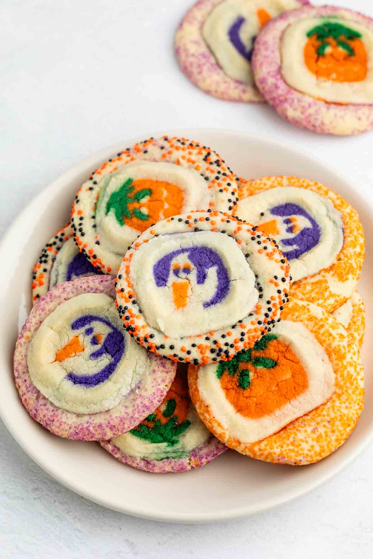 A plate of colorful Halloween cookies decorated with shapes of pumpkins and ghosts, surrounded by festive orange, purple, and black sprinkles.