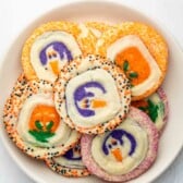A white bowl filled with colorful Halloween-themed sugar cookies decorated with designs like pumpkins and ghosts, edged with orange, purple, and black sprinkles.