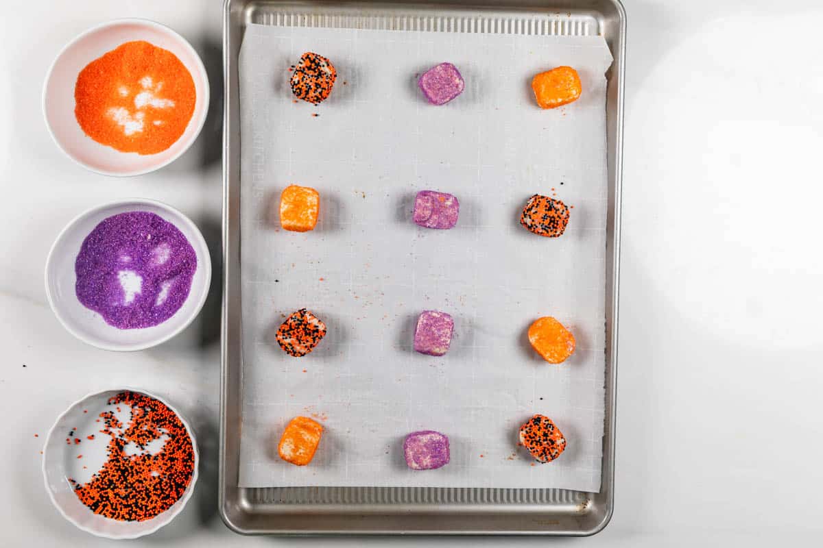A baking sheet lined with parchment paper holds cubes of cookie dough coated in orange, purple, and black sprinkles. Three bowls with the same colored sprinkles are next to the tray on a white surface.