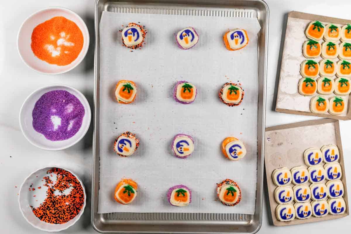 A baking tray with Halloween-themed cookies, decorated with ghost and pumpkin designs, sits on parchment paper. Surrounding it are bowls of orange, purple, and black sprinkles, with more cookies on nearby trays.