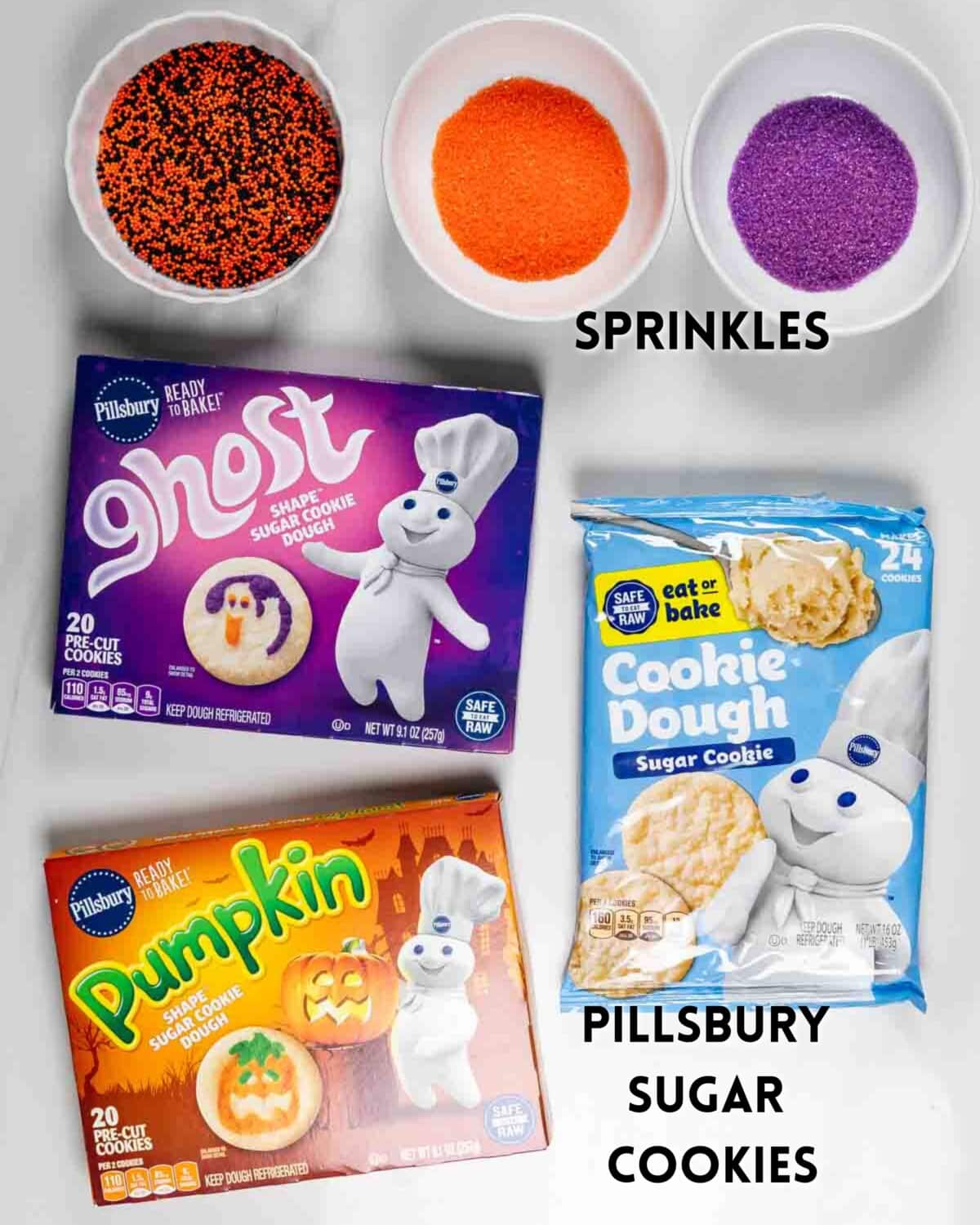 Three bowls of orange, black, and purple sprinkles above three packages of Pillsbury sugar cookies: Ghost shape, Pumpkin shape, and plain cookie dough, arranged on a white surface.
