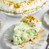 A slice of creamy green pistachio pie topped with whipped cream and chopped pistachios sits on a white plate, with the rest of the pie and utensils visible in the background.