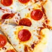 Slices of pepperoni pizza with melted cheese and tomato sauce on a light-colored surface. A small bowl with additional pepperoni slices is visible in the background.