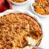 A round baking dish filled with pumpkin crisp topped with a golden crumbly streusel, a large serving scooped out. Nearby are two small bowls of the dessert, one with a scoop of vanilla ice cream.