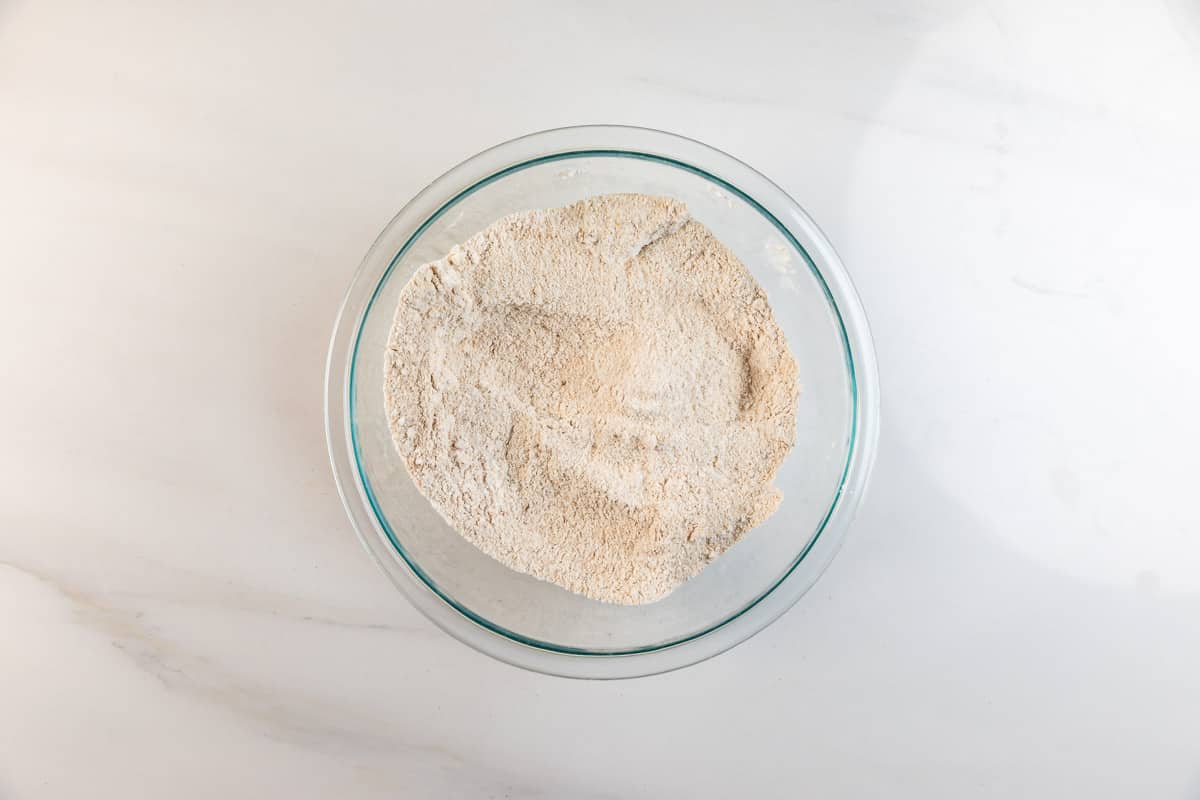 A clear glass bowl filled with a mound of light brown flour mixture sits on a white marble surface.