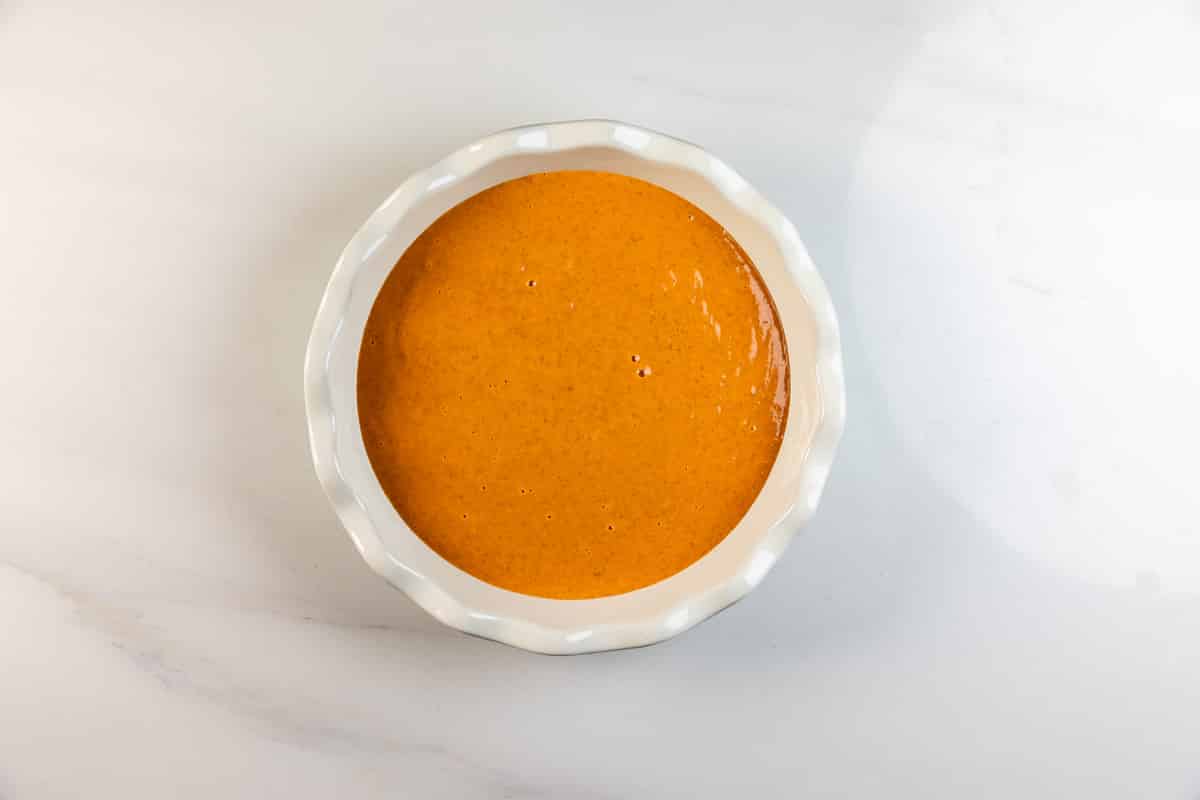 A white ceramic dish filled with smooth, orange-colored sauce or batter, placed on a white marble surface.