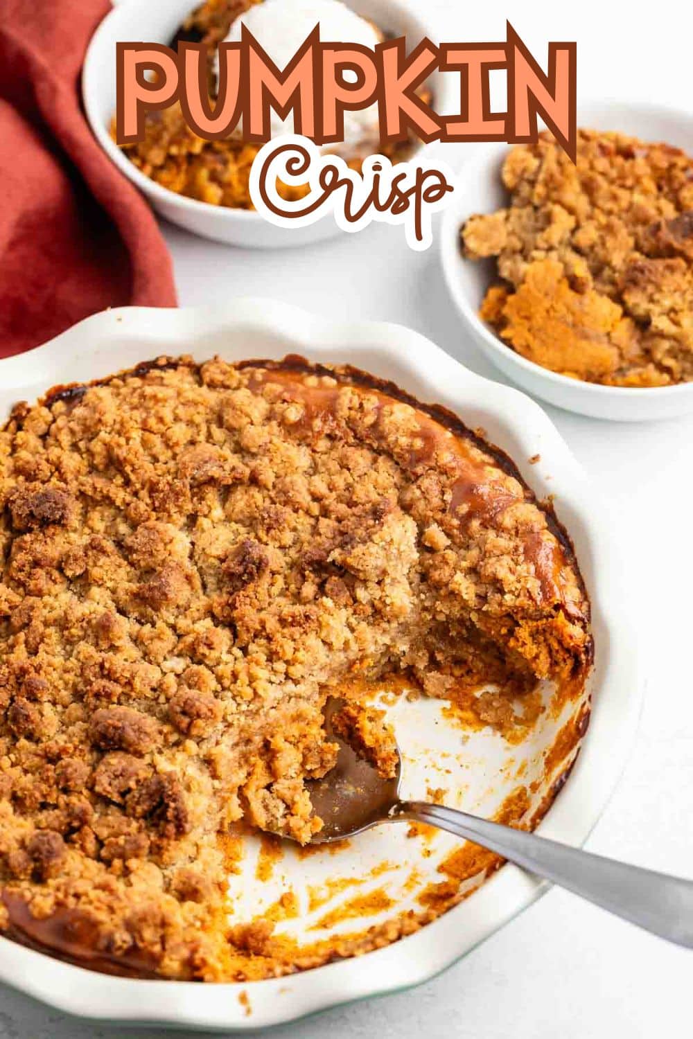 A white pie dish filled with pumpkin crisp topped with a crumbly brown streusel, with a serving missing. A spoon rests in the dish. Two small bowls with portions of pumpkin crisp are in the background. Text reads Pumpkin Crisp.
