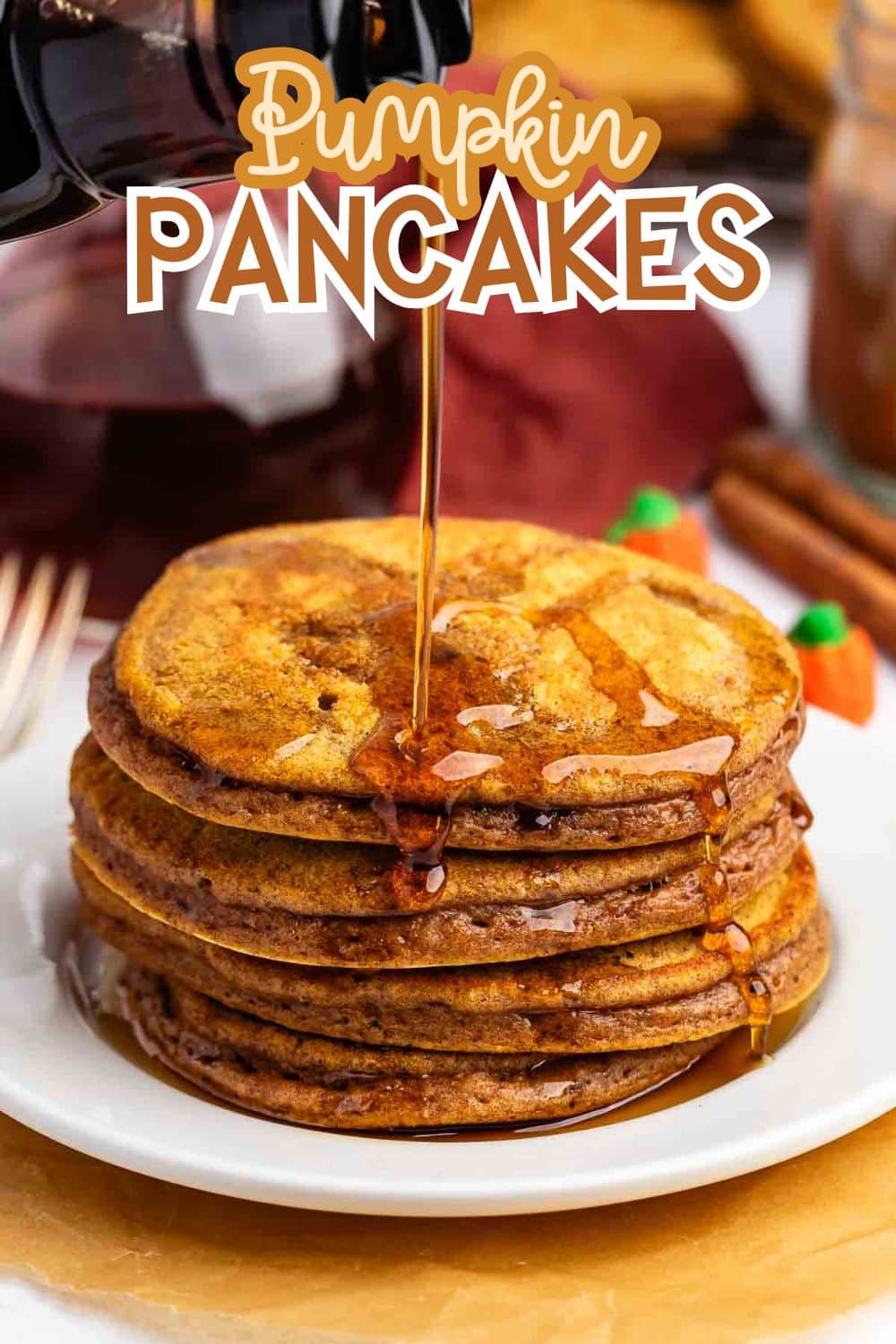 A stack of pumpkin pancakes on a white plate is being drizzled with syrup. The words Pumpkin Pancakes are written above the stack, and some cinnamon sticks and a fork are in the background.