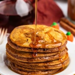 A stack of pumpkin pancakes on a white plate is being drizzled with syrup.