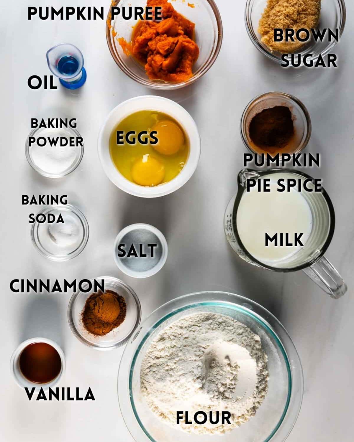 Various baking ingredients are arranged on a white surface, each labeled: pumpkin puree, brown sugar, oil, baking powder, baking soda, eggs, pumpkin pie spice, milk, cinnamon, salt, vanilla, and flour.