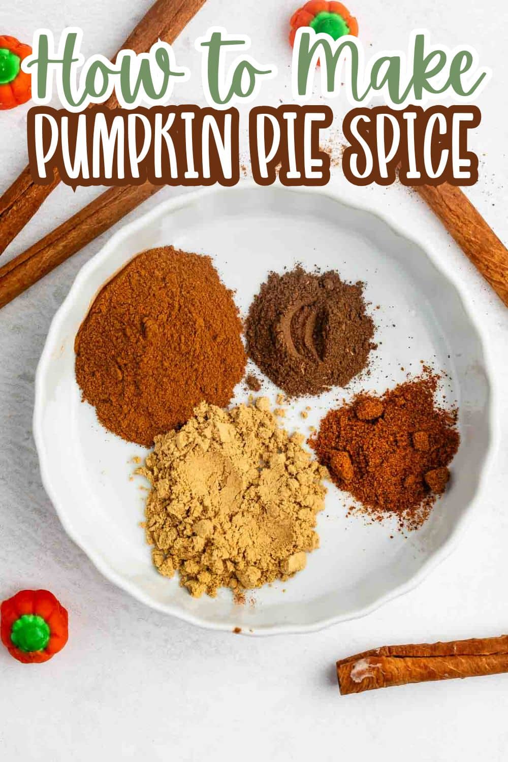 A white plate holds four piles of ground spices: cinnamon, nutmeg, ginger, and allspice, with cinnamon sticks and candy pumpkins nearby. Text reads How to Make Pumpkin Pie Spice.
