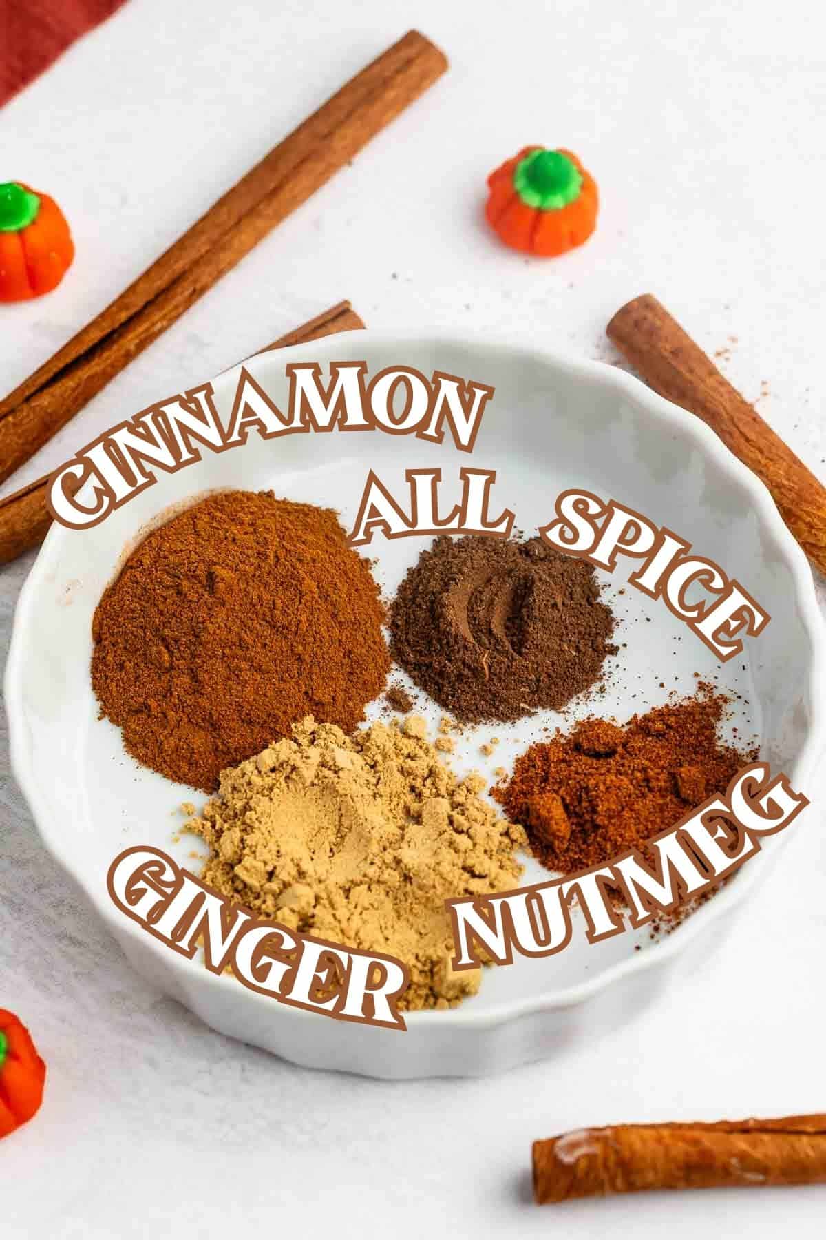 A white dish holds four piles of ground spices labeled as cinnamon, allspice, ginger, and nutmeg—key ingredients in pumpkin pie spice. Cinnamon sticks and small pumpkin-shaped candies are arranged around the dish on a white surface.