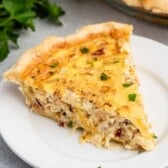 A slice of quiche on a white plate. The quiche has a golden crust and is filled with a creamy mixture containing small bits of bacon and herbs. Fresh parsley is scattered on top and around the plate.