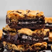 Three stacked dessert bars with layers of chocolate chip cookie dough, Oreos, and brownie, showing gooey chocolate and cookie texture, with more dessert bars and Oreos in the background.